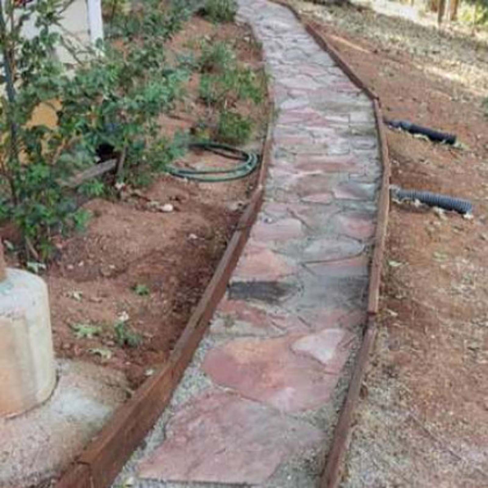 A flagstone pathway edged with brown timber planks winds through an outdoor dirt area next to a building.