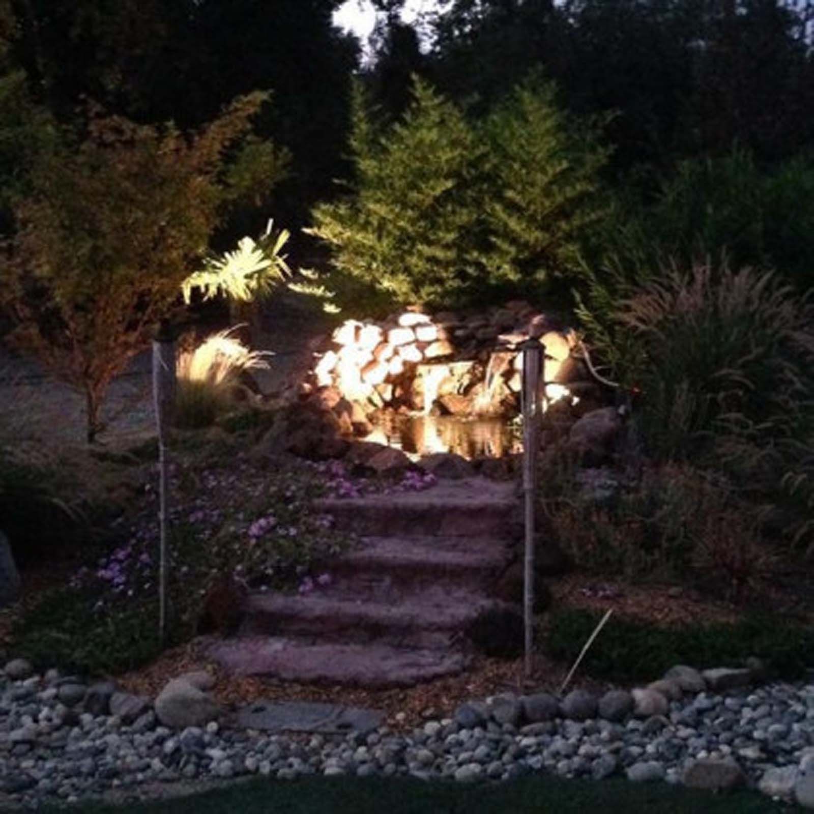 A small, illuminated backyard pond with a stone waterfall at the top of a short set of stairs, surrounded by landscaping.
