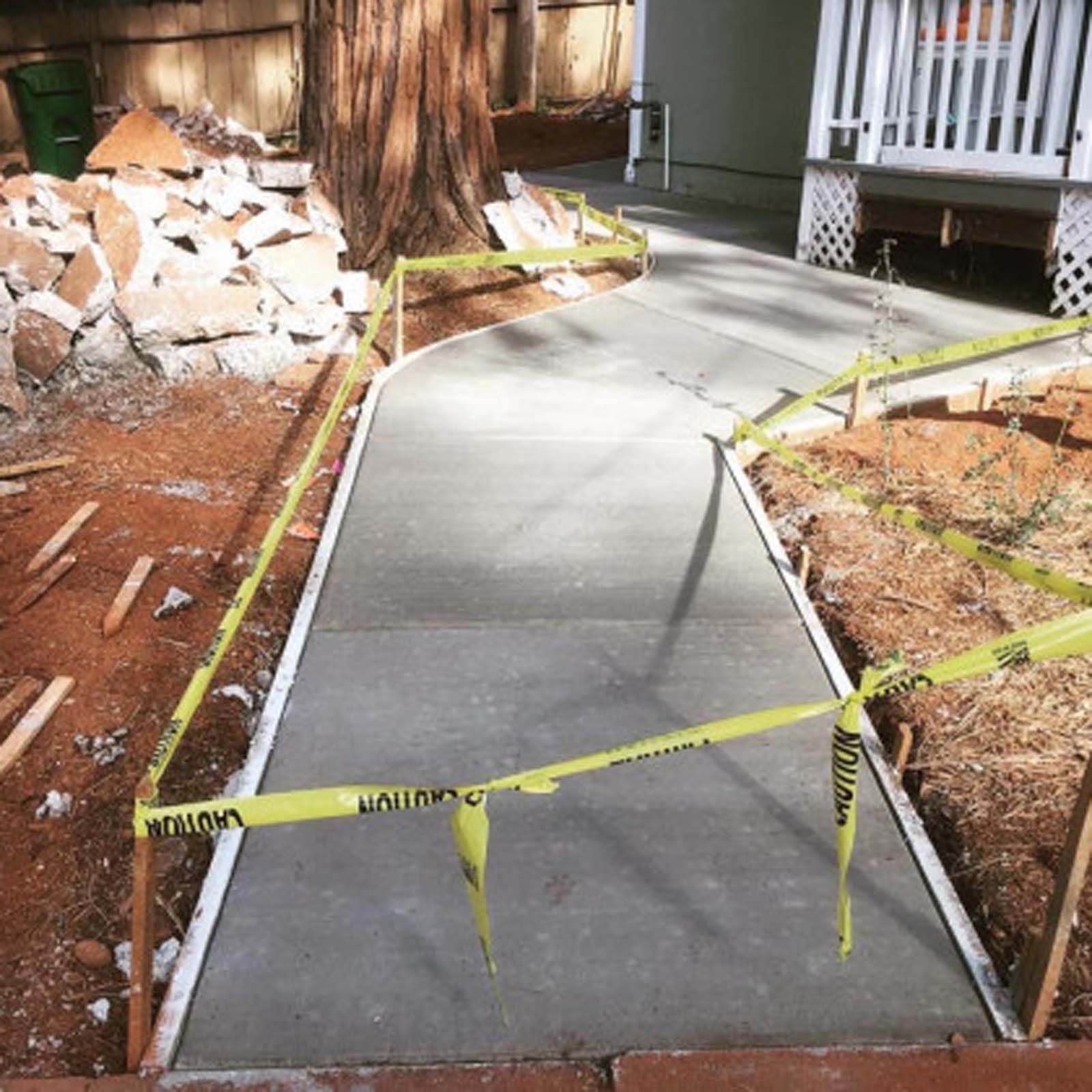 Newly poured concrete walkway surrounded by yellow caution tape, leading to a porch next to a large tree.