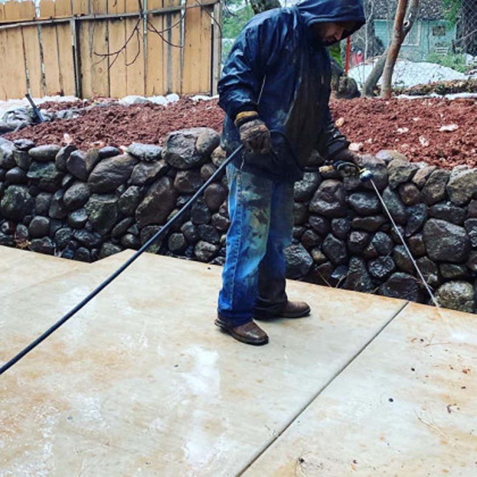 A person in a hooded jacket and work clothes pressure washes a concrete patio in front of a stone retaining wall.