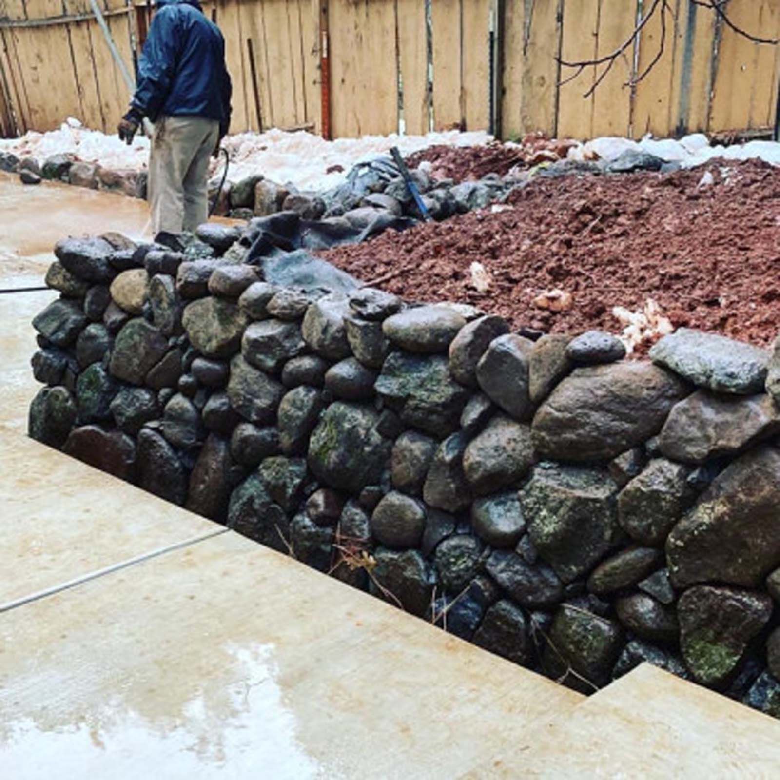 A person stands near a newly constructed stone retaining wall next to a bed of dark soil and a concrete patio.