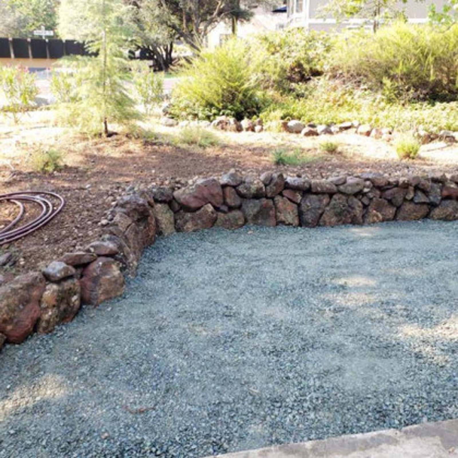 A low, dry-stacked stone wall curves along the edge of a gravel-covered patio in a sunny, landscaped backyard.