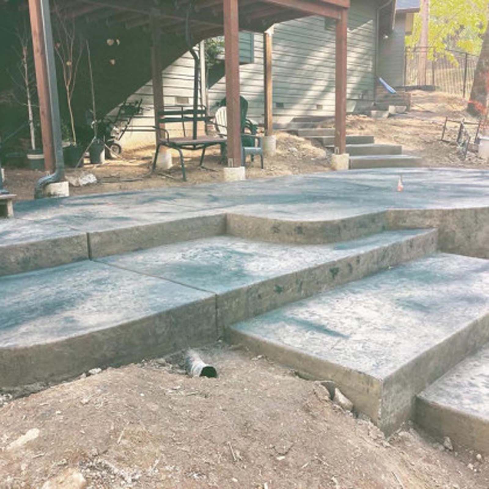 A backyard with a newly installed gray stamped concrete patio featuring wide, tiered steps leading to a wooden deck.