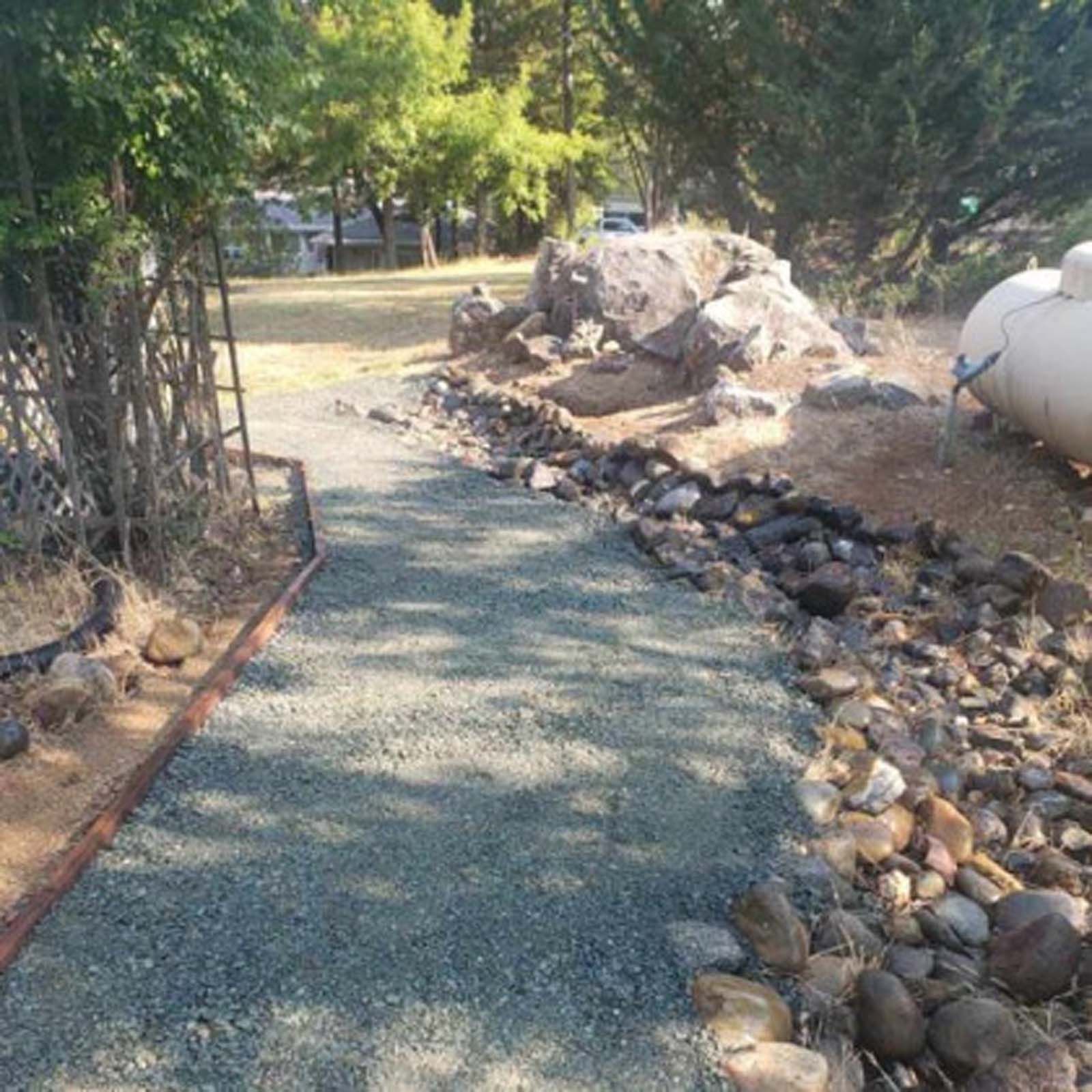 A gravel path bordered by metal edging and rocks leads toward large boulders and a propane tank in a sunny yard.