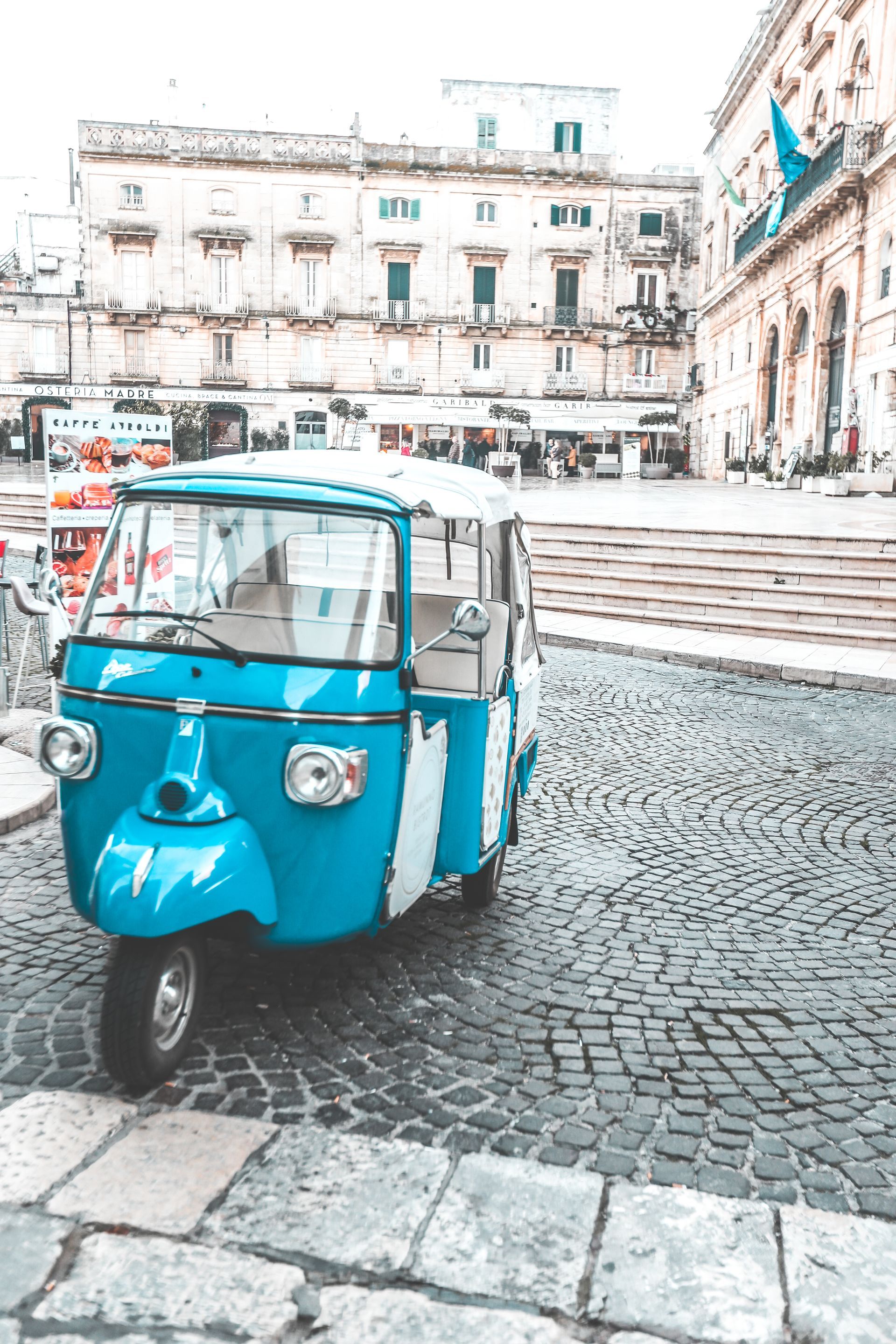A car parked on three wheels in a square in Ostuni, with a picturesque view of the city's white-washed buildings in the background.