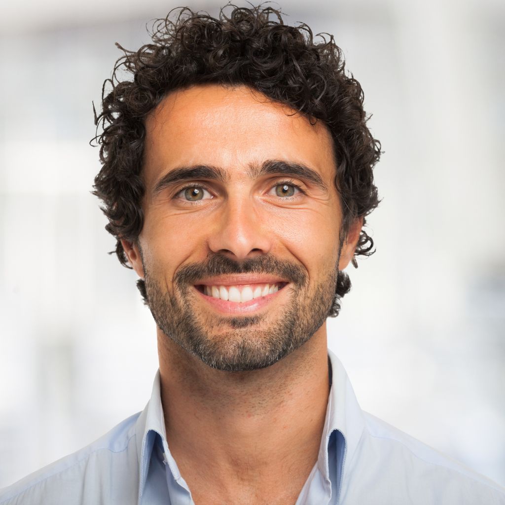 Man with curly dark hair and beard smiles, blue shirt, blurred background.
