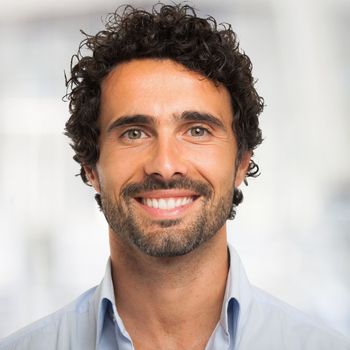 Man with curly dark hair and beard smiles, blue shirt, blurred background.