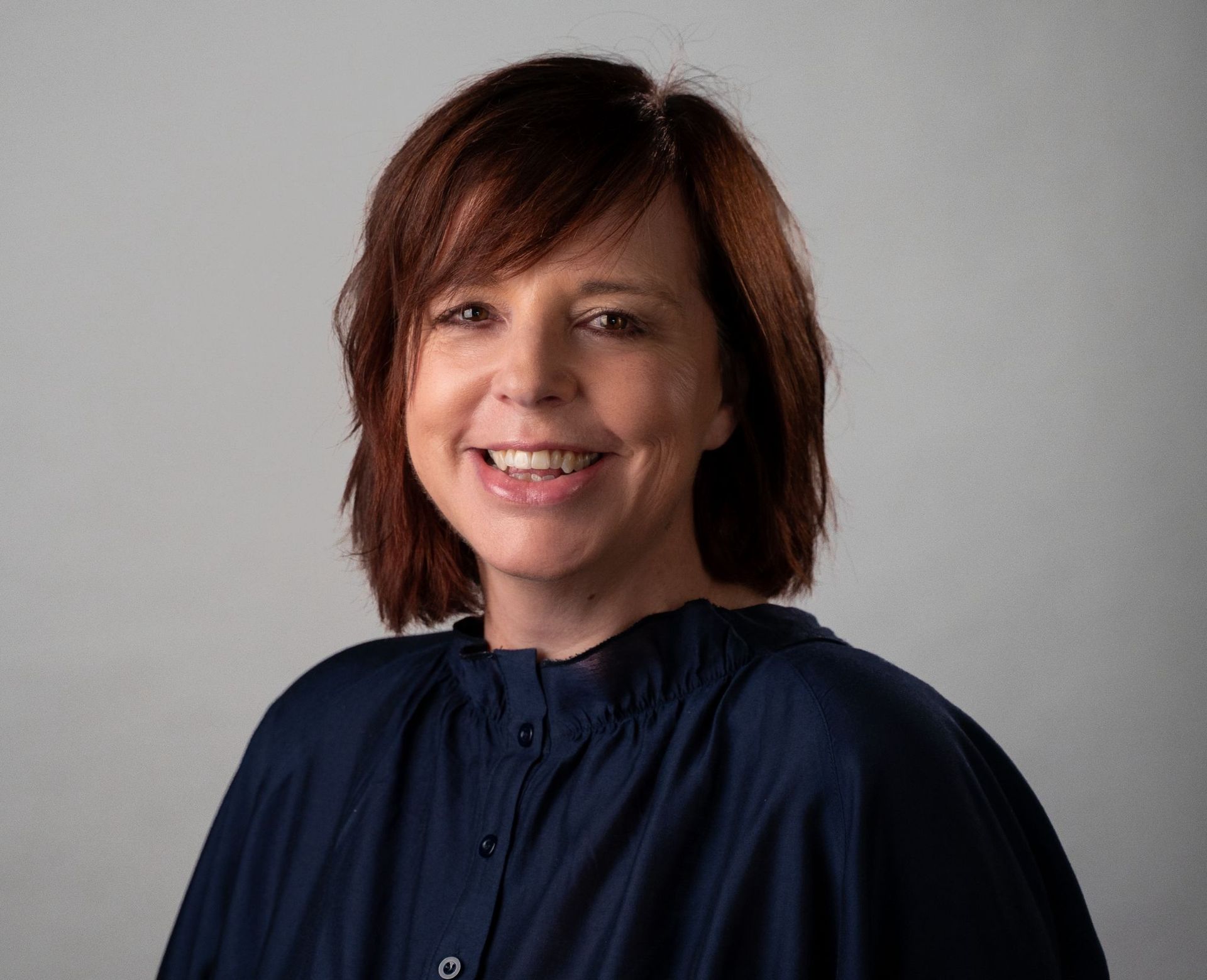 Woman with red-brown hair smiles, wearing a dark blue blouse against a gray background.