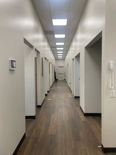 Long, empty hallway with white walls, brown doors, and wood-look flooring. Fluorescent ceiling lights.