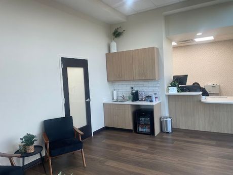 Modern waiting room with a reception desk and coffee station. Includes a black chair, plants, and wooden floors.