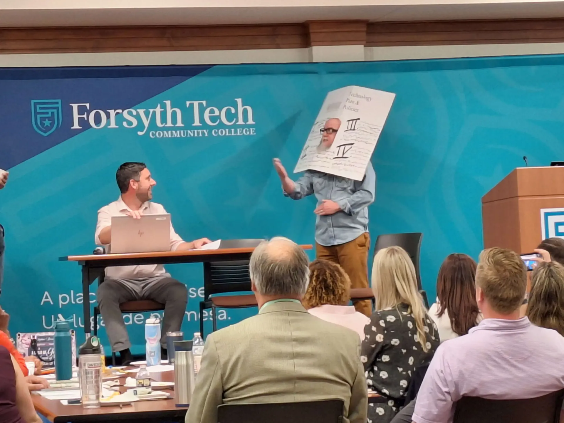 A person wearing a large paper cutout of a face over their head stands before an audience at a Forsyth Tech event.