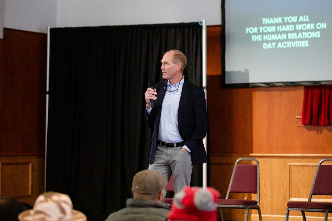A person in a blazer and button-down shirt speaks into a microphone in front of a screen displaying thank you text.