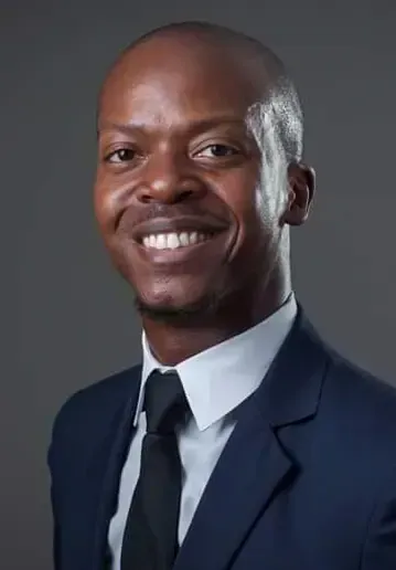 A person in a blue suit, white shirt, and black tie smiling at the camera against a plain dark gray background.