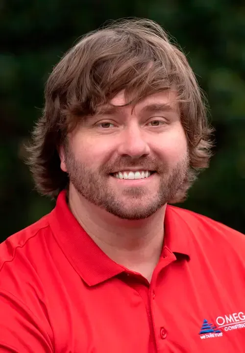 A smiling person with shoulder-length brown hair and a beard, wearing a red polo shirt, against a blurred green background.