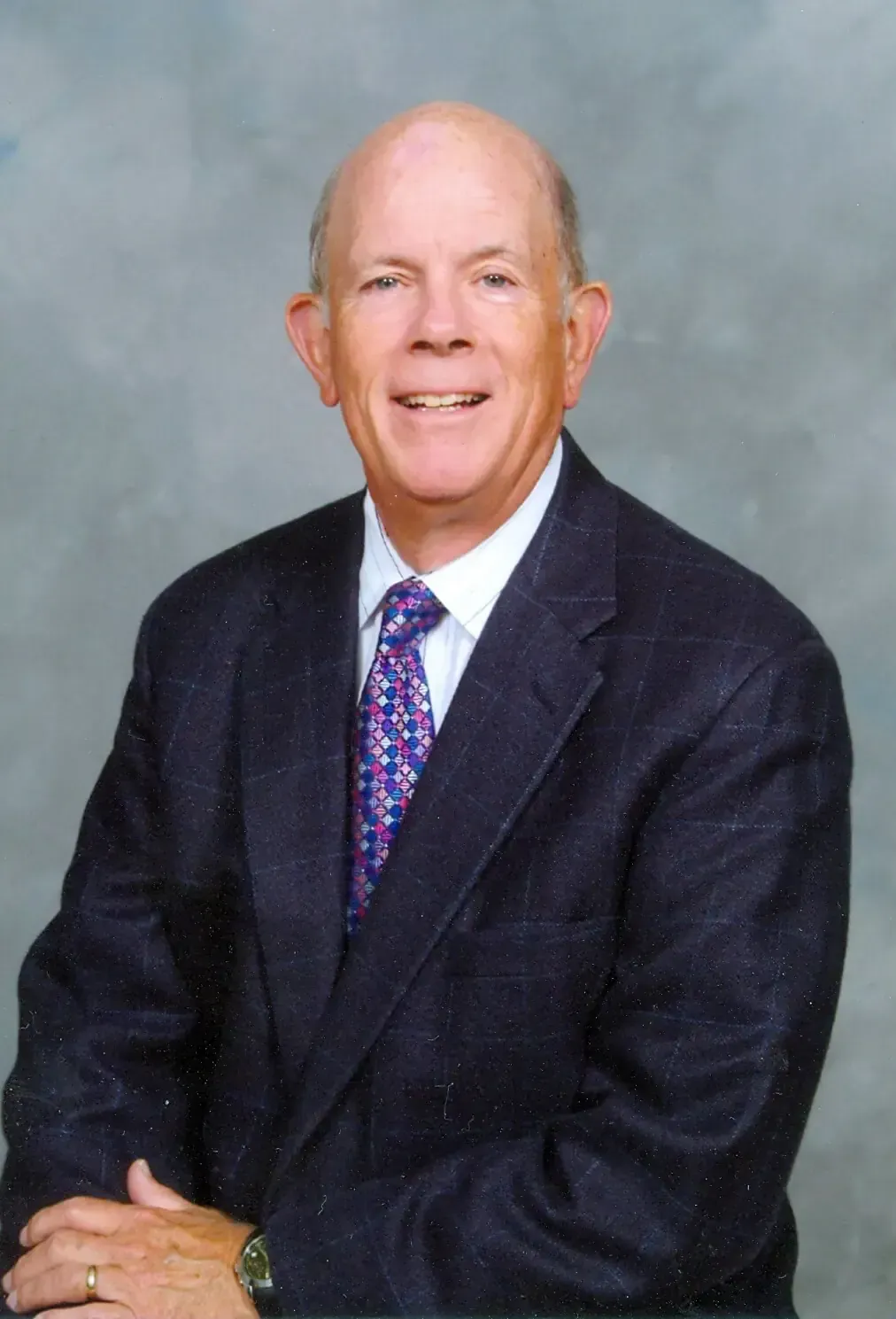 A smiling person wearing a dark blue blazer, white collared shirt, and patterned purple tie against a grey background.