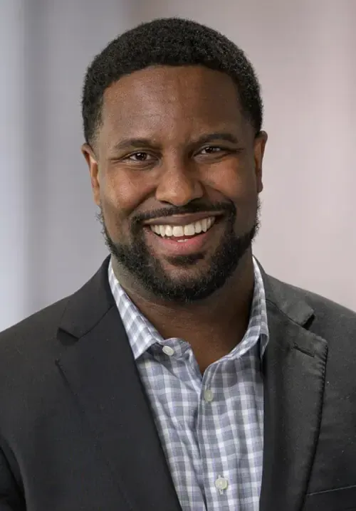 A smiling man in a dark suit jacket and a light-colored checkered dress shirt against a blurred background.