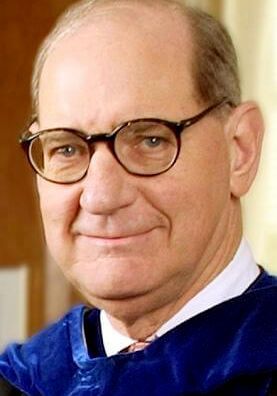 A person wearing round, dark-rimmed glasses and blue academic regalia, smiling slightly against a soft, blurred background.
