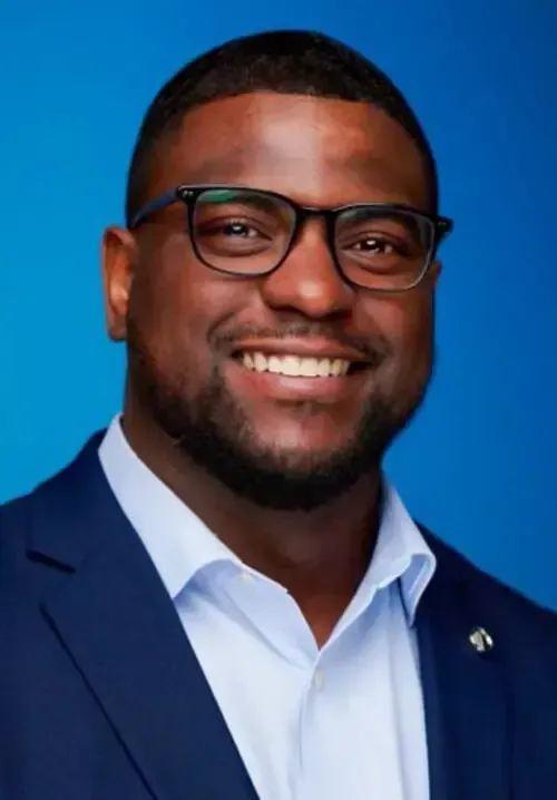 A person with a beard and glasses, smiling in a blue shirt and suit jacket, against a bright blue background.