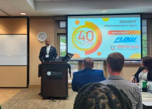 A person speaks at a podium in a conference room before an audience, with a presentation screen displaying