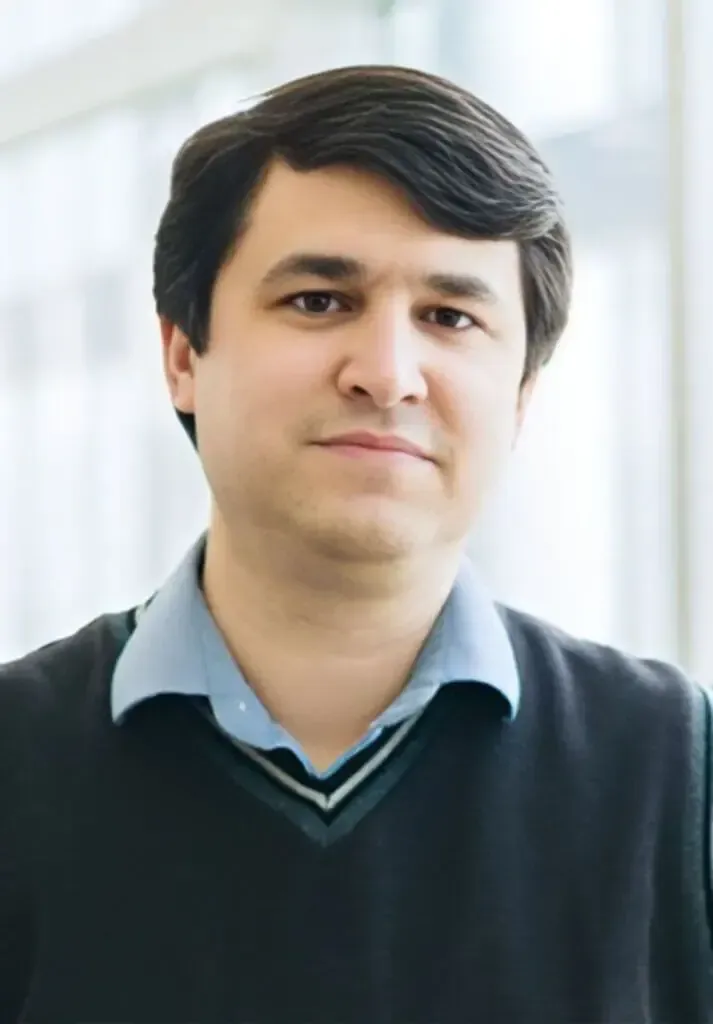 Headshot of a person with dark hair, wearing a collared blue shirt under a dark V-neck sweater, standing before a window.