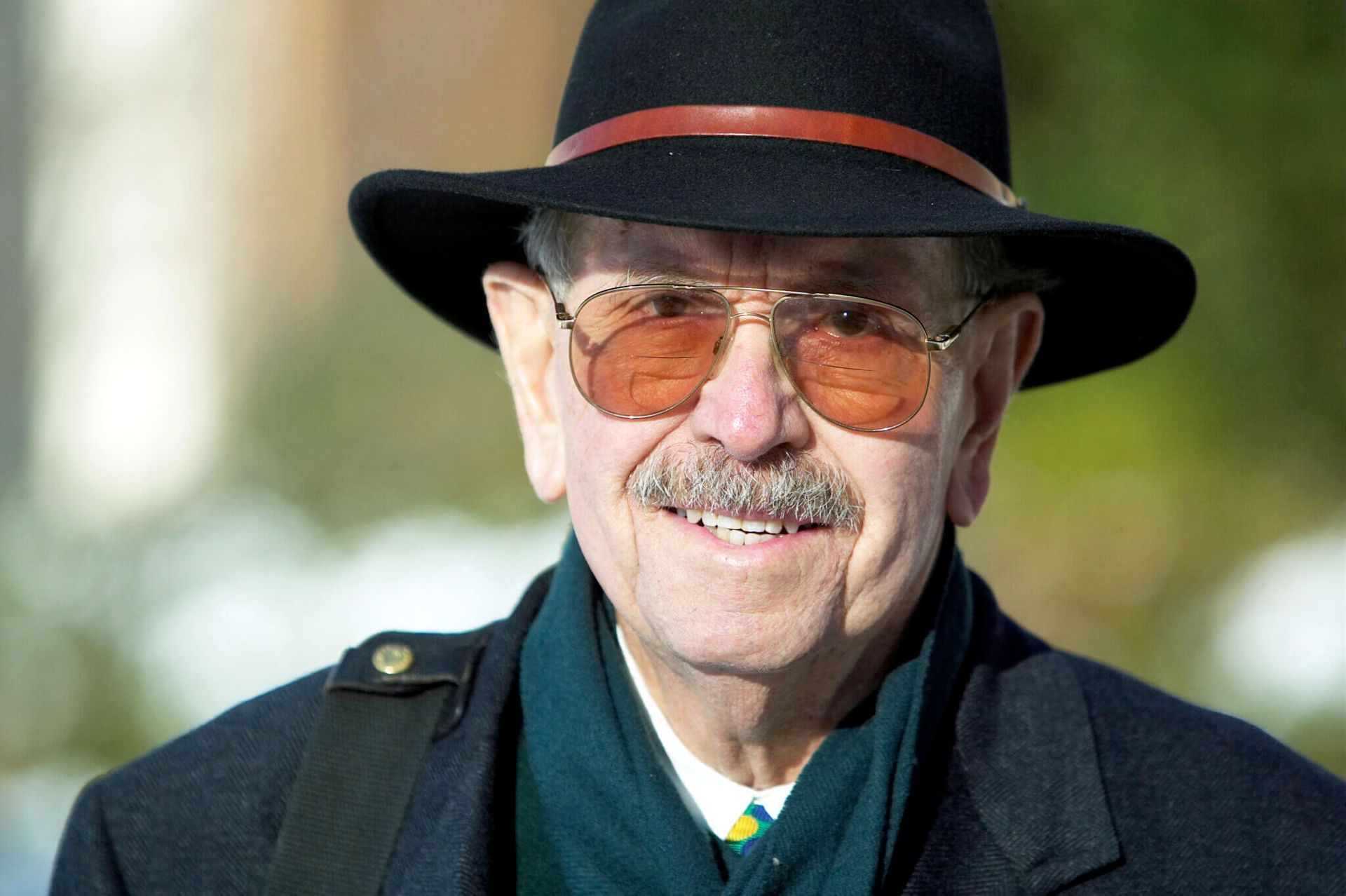 A person smiling while wearing tinted glasses, a green scarf, a dark coat, and a fedora with a brown band.