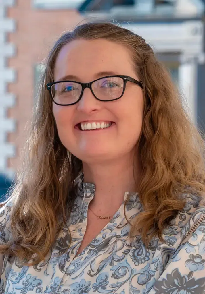 Smiling person with long wavy hair and glasses wearing a blue and white floral shirt, standing outdoors.