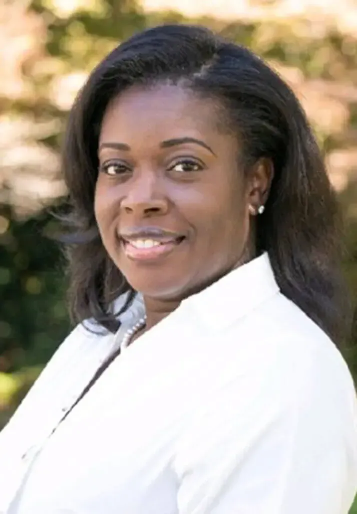 A smiling person with dark hair, wearing a white collared shirt, posing for a portrait in a sunlit, outdoor setting.