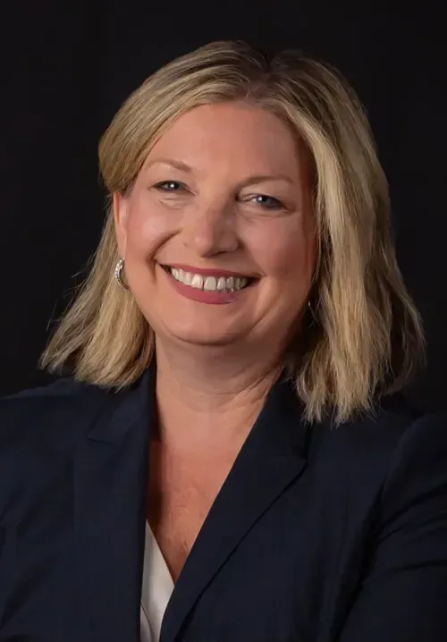 A smiling professional wearing a navy blazer and white top, centered against a dark background.