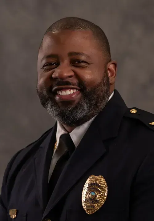 A smiling man in a dark suit jacket and a light-colored checkered dress shirt against a blurred background.