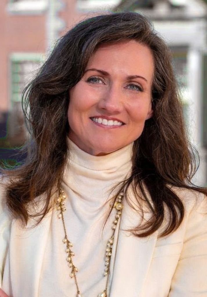Smiling person with long, dark hair, wearing a cream-colored turtleneck and blazer with a gold necklace, outdoors.