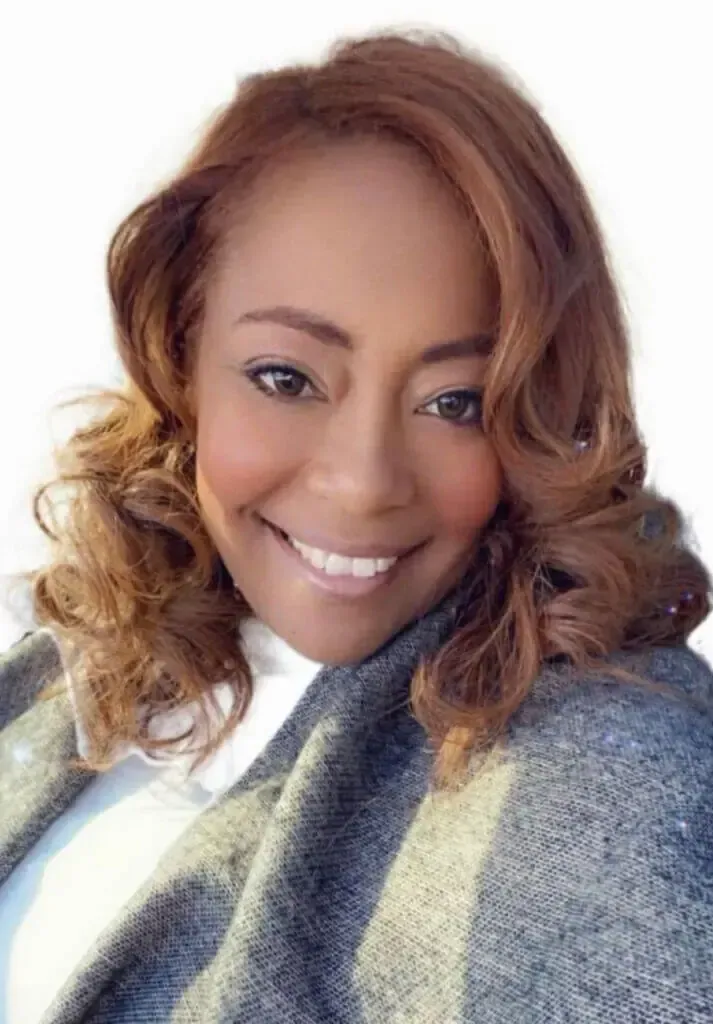A smiling person with shoulder-length, light brown wavy hair wearing a white top and a grey textured scarf.