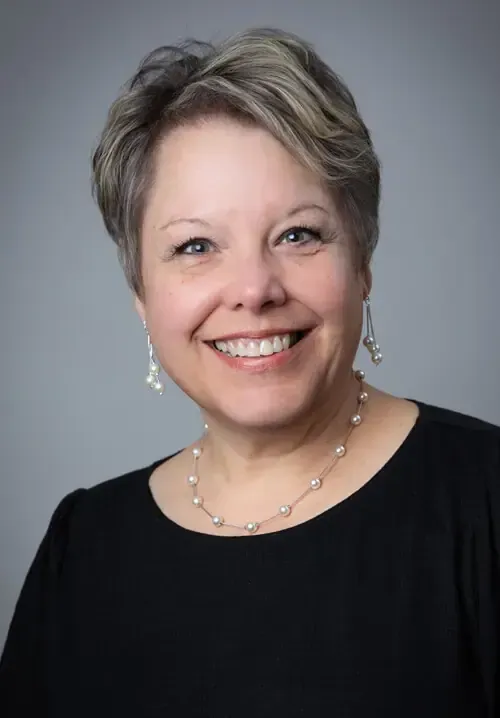 Headshot of a smiling person with short, light hair wearing a black top and a pearl necklace against a gray background.