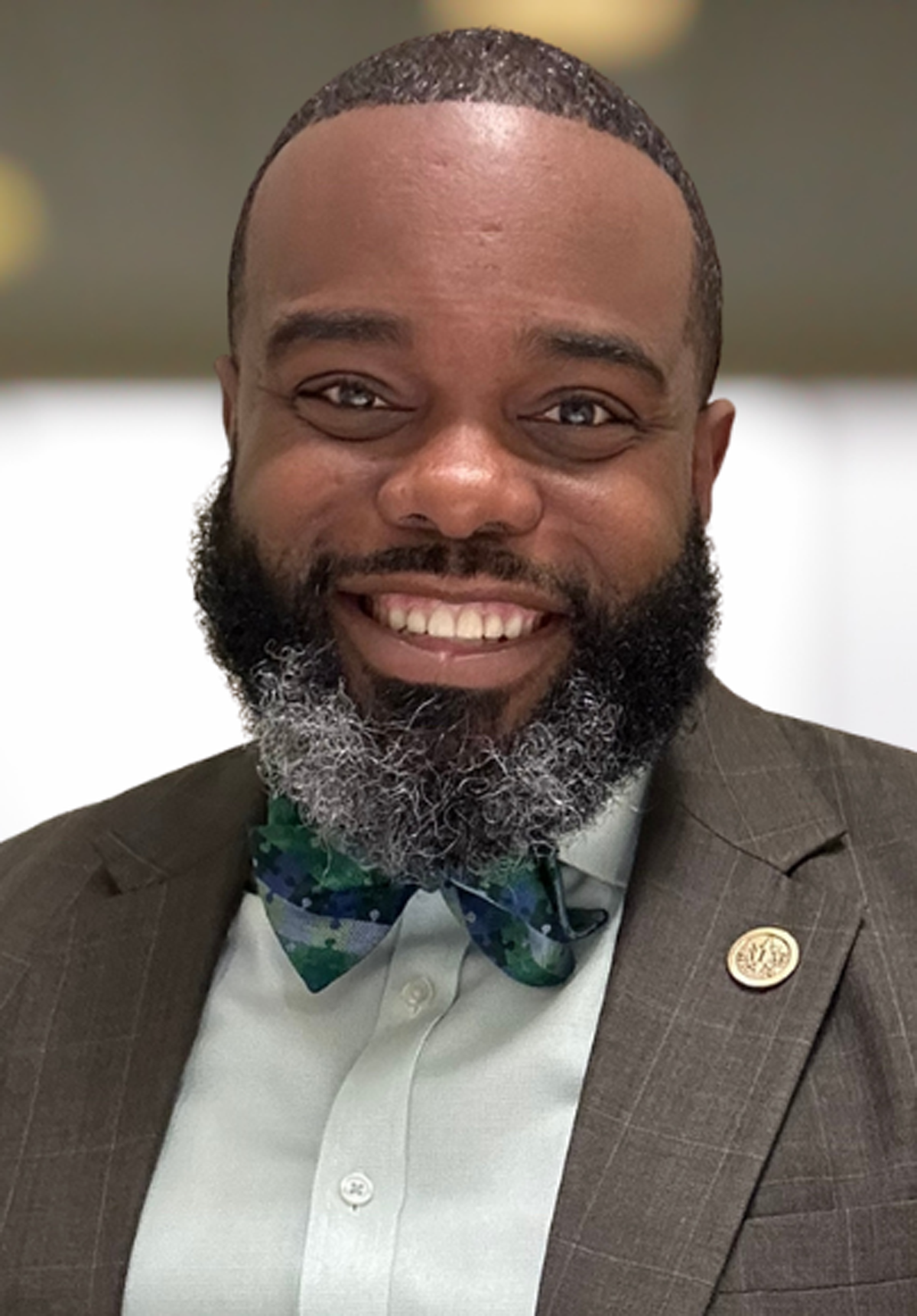 Smiling man in a brown suit and green bow tie with a lapel pin, against a blurred indoor background
