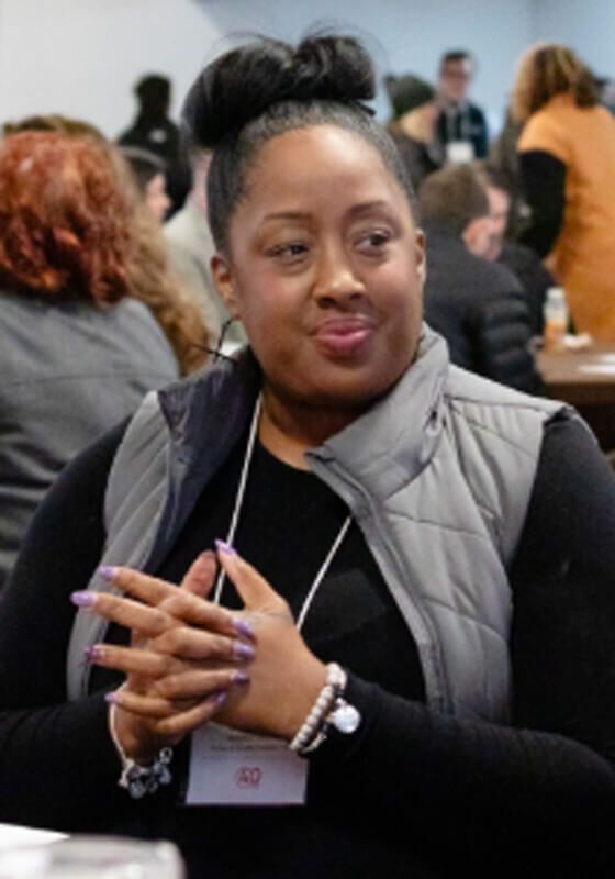 A person with hair in a bun wears a black top and gray vest, smiling while seated with their hands clasped at a table.