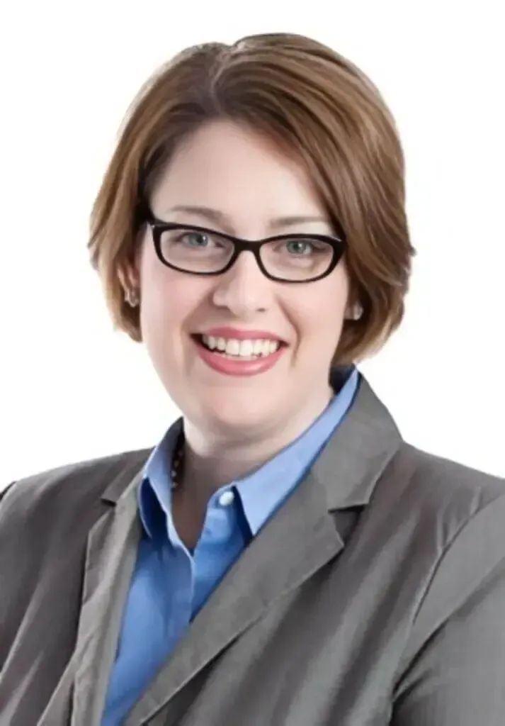 Headshot of a person with short brown hair and glasses, wearing a grey blazer and blue shirt, smiling against white.