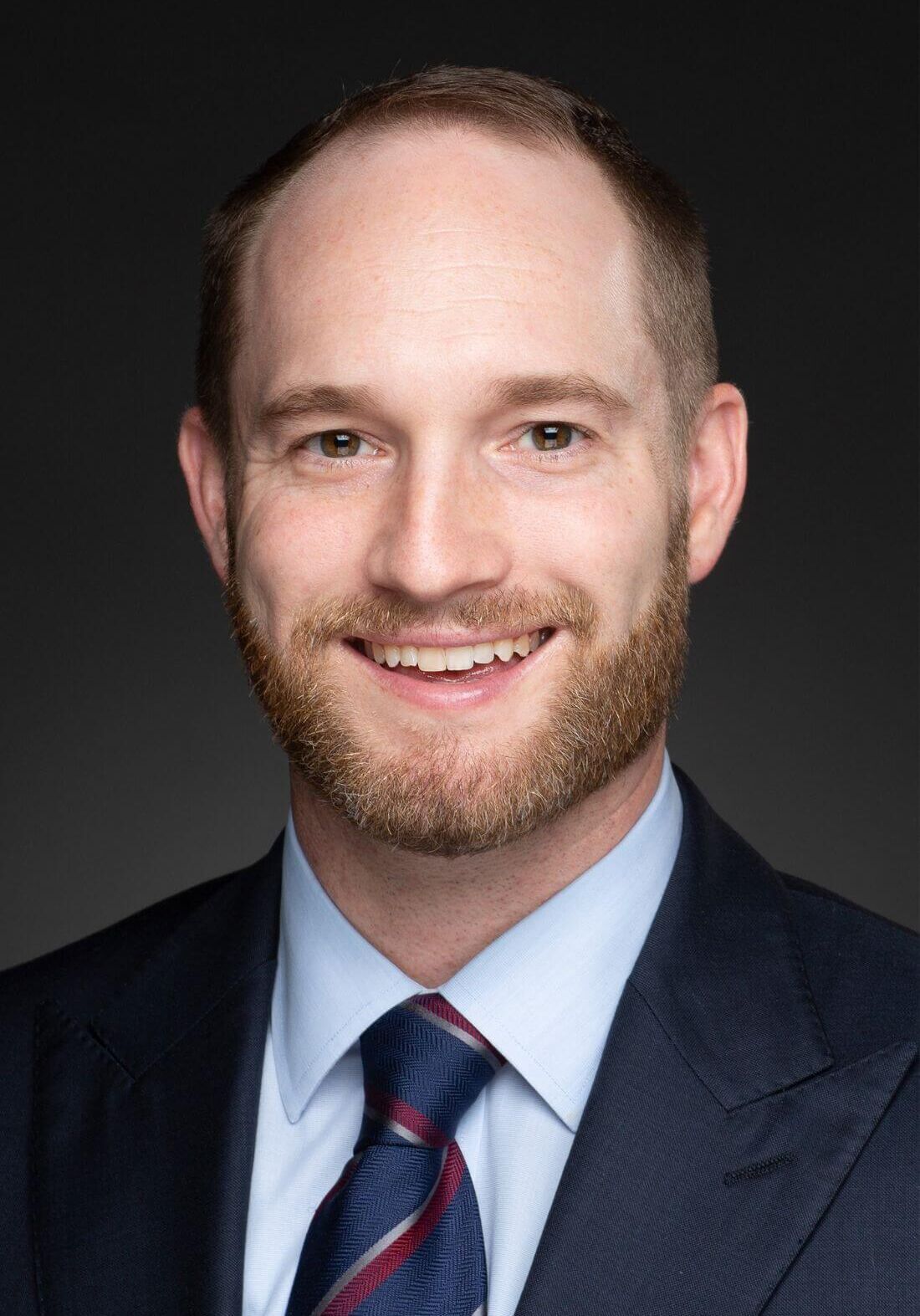 Headshot of a smiling person with a beard, wearing a dark suit, light blue dress shirt, and a blue patterned tie.