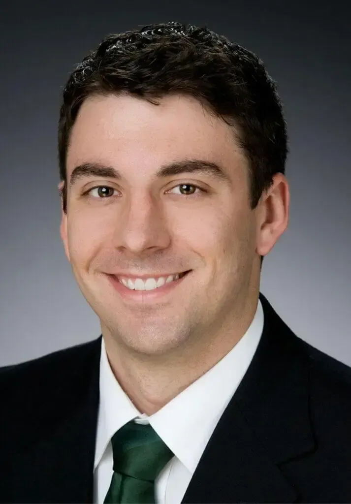 Headshot of a smiling person wearing a dark suit, white shirt, and green tie against a neutral gray background.