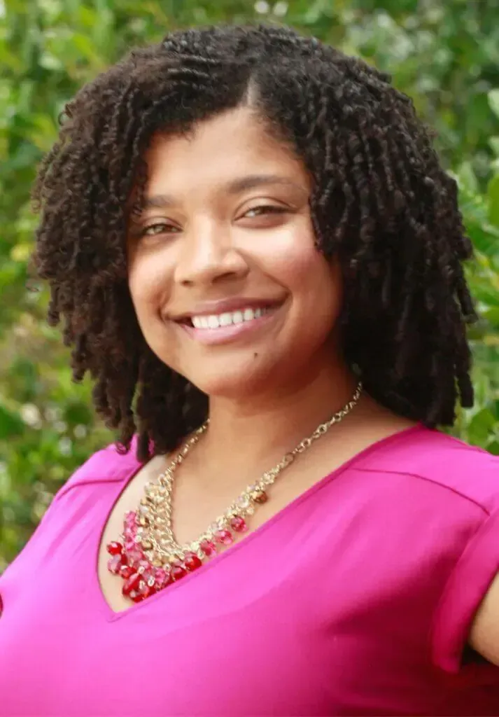 A smiling person wearing a pink top and a layered, multi-toned red and gold beaded necklace, framed by green foliage.