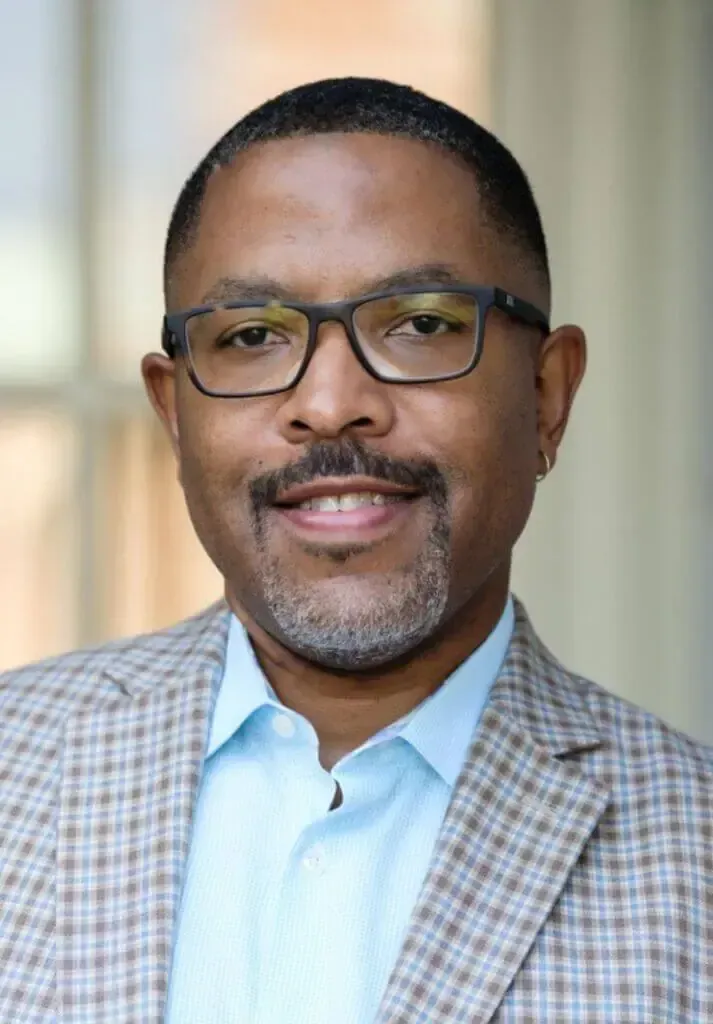 A person with a beard and glasses, smiling in a blue shirt and suit jacket, against a bright blue background.