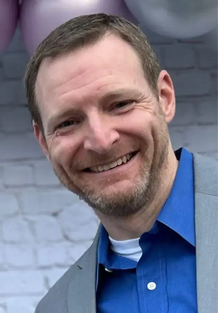 A man with short brown hair and light stubble wearing a blue button-down shirt and grey blazer, smiling at the camera.