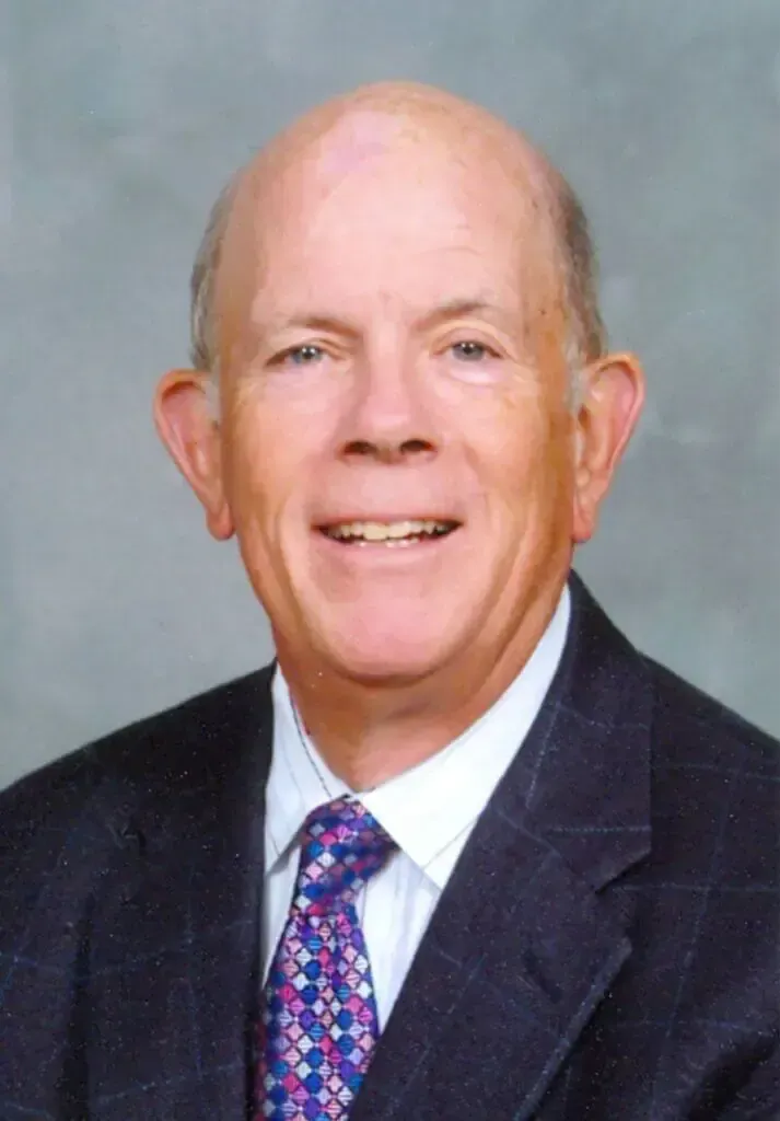 A smiling person in a dark suit with a checkered pattern and a blue and red patterned tie against a grey background.