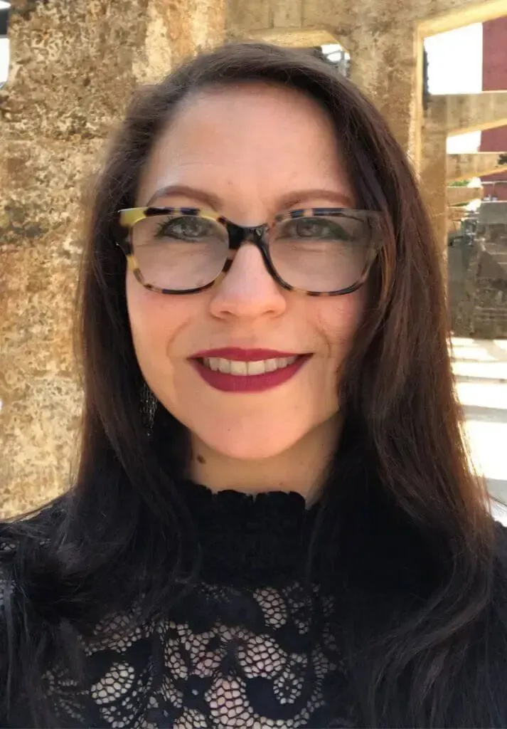 A person with long dark hair and dark-rimmed glasses smiling, wearing a black lace top against a stone wall background.