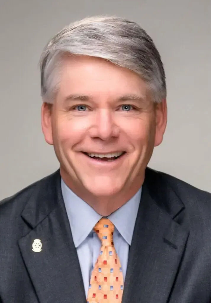 Headshot of a smiling person wearing a dark blue suit, light blue dress shirt, and a patterned orange tie.