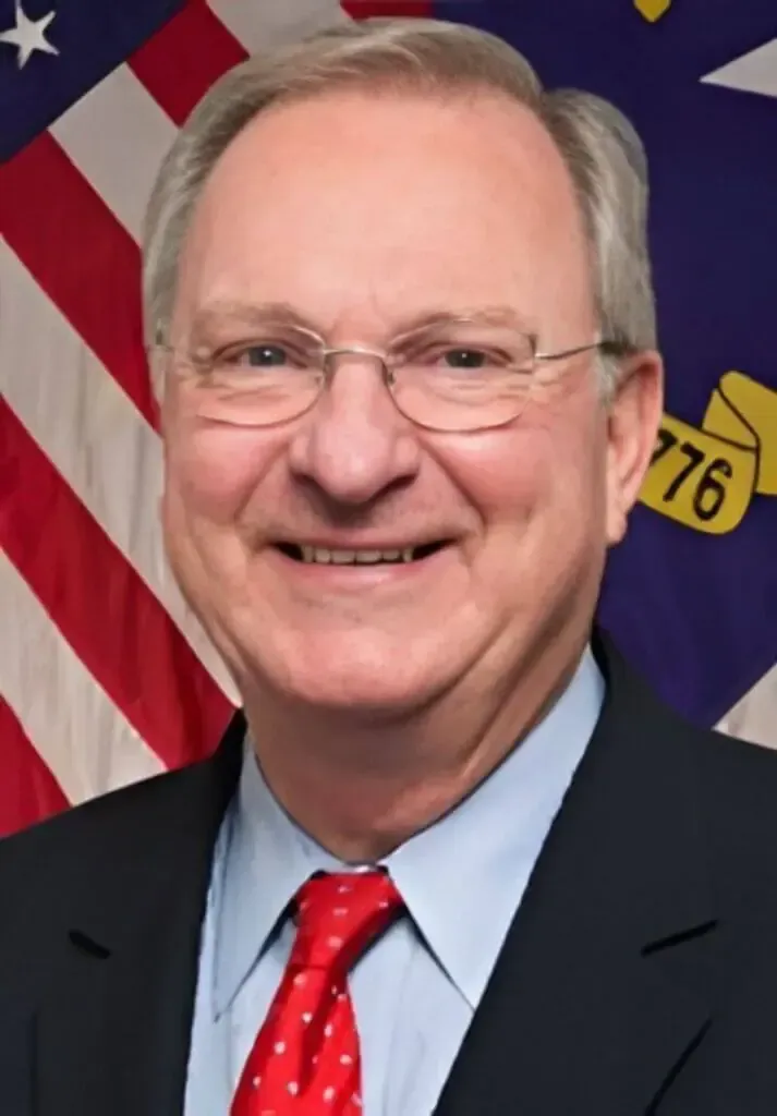 A man in a suit and red patterned tie smiling in front of an American flag and a banner with 