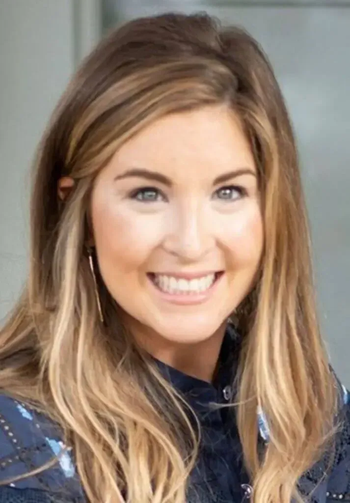 A smiling person with long, blonde-highlighted brown hair wearing a dark, patterned collared shirt.