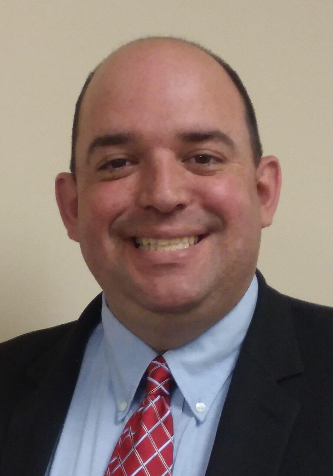 A smiling person in a suit, light blue dress shirt, and red patterned tie against a light-colored wall.