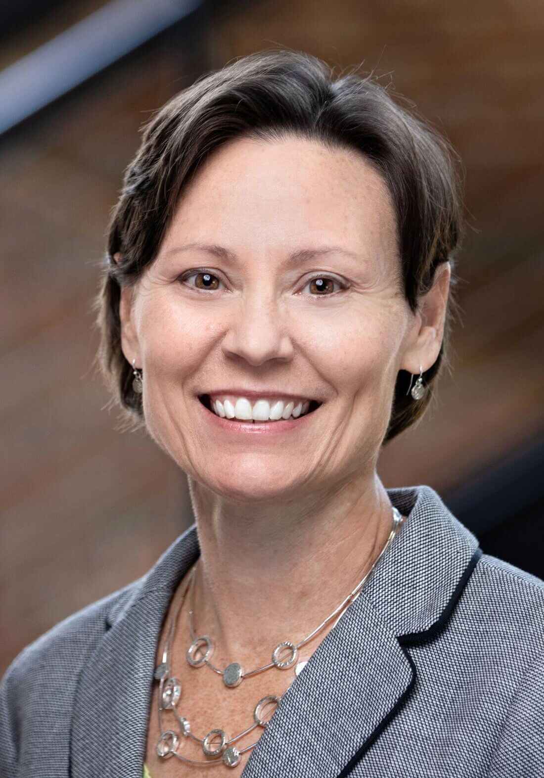 A smiling professional wearing a tan blazer and a silver chain necklace, captured in a headshot against a window.