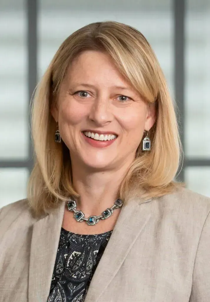 A smiling professional wearing a tan blazer and a silver chain necklace, captured in a headshot against a window.