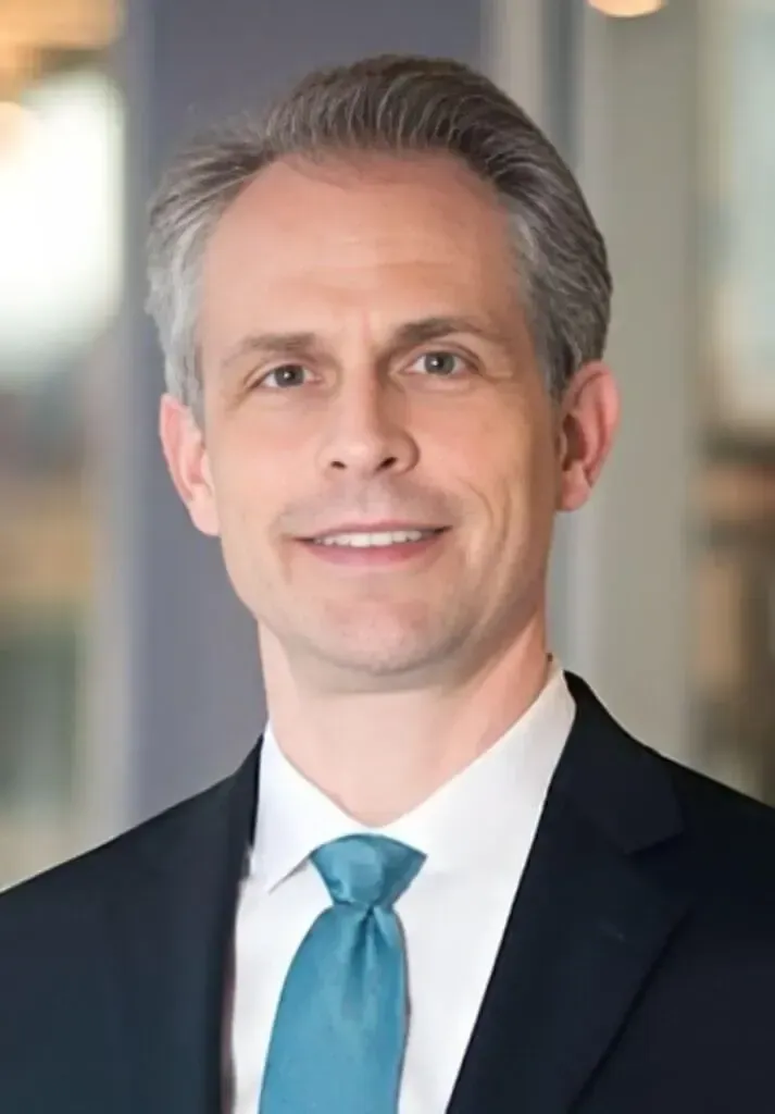 Professional headshot of a person wearing a dark suit, white shirt, and teal tie, centered against a blurred background.