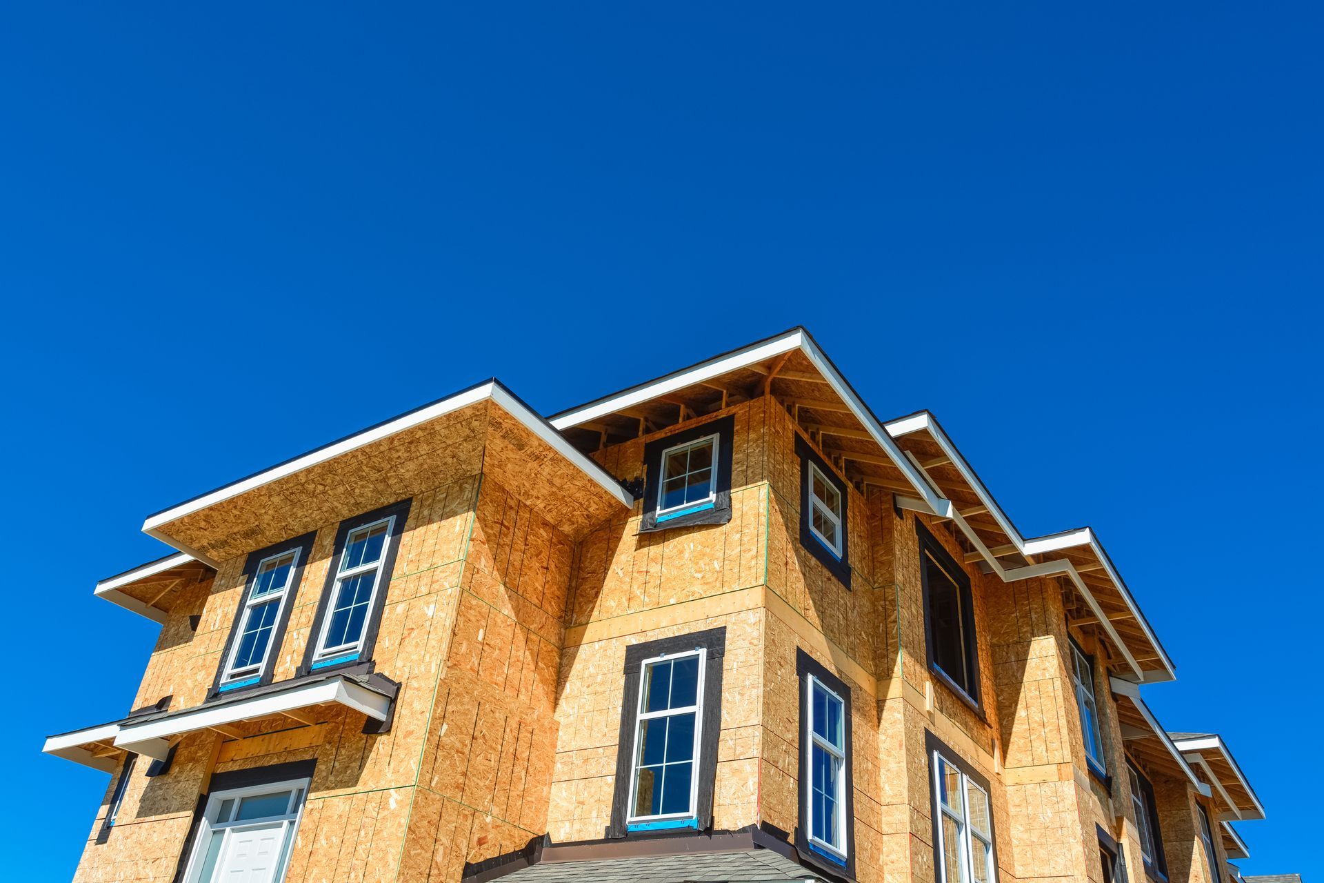 Une maison à plusieurs étages en construction, avec des panneaux de bois apparents, des fenêtres encadrées et un ciel bleu clair en arrière-plan.